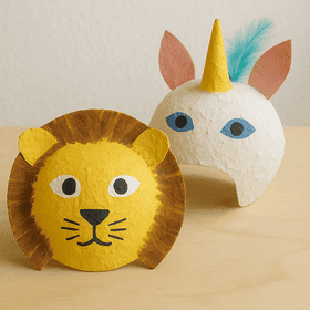 Zwei selbstgebastelte Pappmaché-Masken – ein gelber Löwe mit Mähne und ein weißes Einhorn mit Horn und Federn – auf Holztisch.