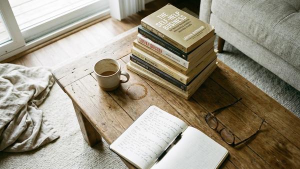 Ein Holztisch mit einem Stapel Bücher, einer Kaffeetasse mit Fleck und einem offenen Notizbuch, das eine gemütliche aber leicht chaotische Szene darstellt.