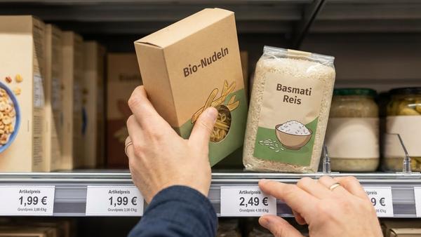 Du siehst eine Hand, die zwei ähnliche Produkte am Regal vergleicht, mit sichtbaren Preisschildern für den Grundpreis.