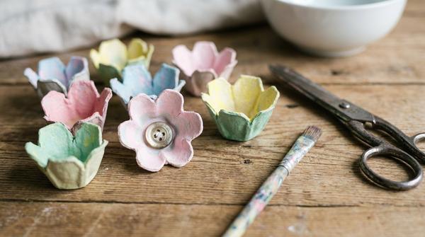 Bunte kleine Blumen aus Eierkarton, bemalt in Pastelltönen, liegen auf einem Basteltisch.