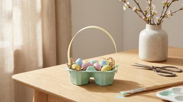 Ausgeschnittenes Eierkarton-Nest als Mini-Osterkörbchen, pastell bemalt und mit Papiergras sowie Schokoeiern gefüllt.