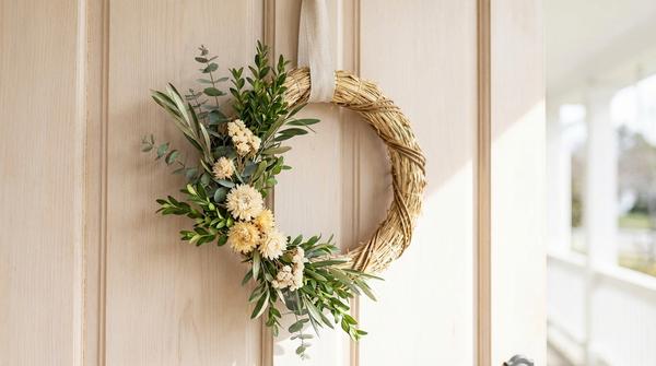 Schlichter Türkranz mit Grünzweigen, kleinen Trockenblumen und Band an einer hellen Tür.