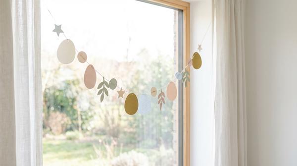 Dezente Papier-Girlande mit Eierformen hängt seitlich an einem hellen Fenster.