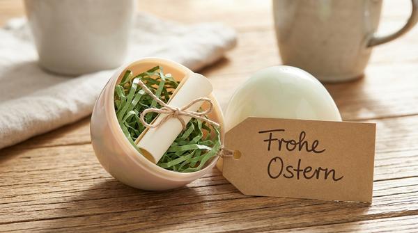 Aufgeklapptes Oster-Ei mit Papiergras und einem eingerollten Gutschein, zusammengebunden mit Band.