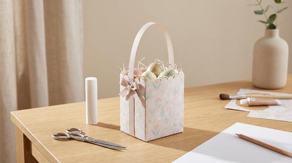 Ein bemalter und beklebter Milchkarton als Osterkörbchen mit Band-Henkel und Papiergras, fotografiert im hellen Tageslicht.