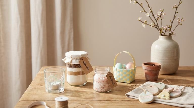 Verschiedene kleine DIY-Ostergeschenke auf einem Holztisch: Glas-Teelicht, Keks-Mix im Glas, Badesalz, Papierkörbchen und mehr.