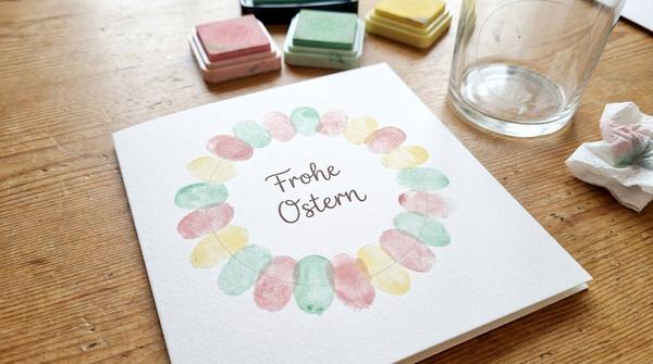 Hände tupfen Pastell-Fingerabdrücke zu einem Blütenkranz auf eine weiße Osterkarte.