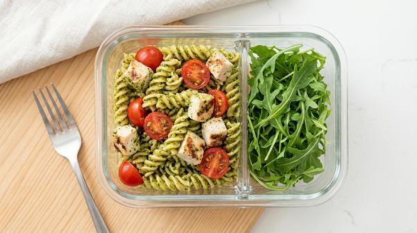 Kalter Pesto-Pasta-Salat mit Hähnchenstücken und Tomaten in einer Lunchbox.