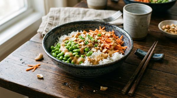 Eine schnelle asiatische Bowl mit Edamame, Reis und cremiger Erdnusssoße auf einem dunklen Holztisch.