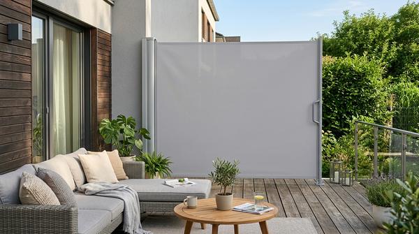Eine ausziehbare Seitenmarkise auf einer modernen Terrasse, die im rechten Winkel von der Hauswand weggezogen ist. An der Kante ist ein Haltegriff im Bodenpfosten eingehakt.
