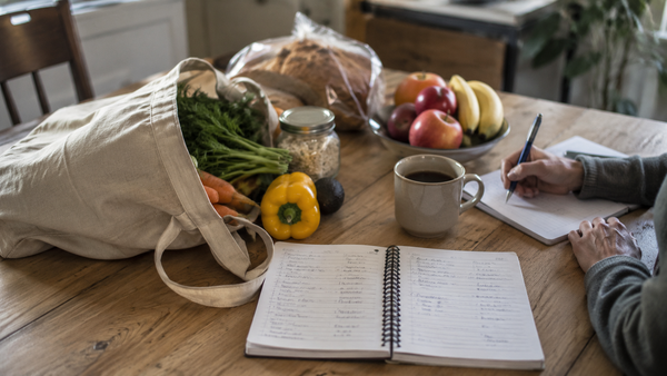 Person schreibt eine Einkaufsliste neben frischen Lebensmitteln, Brot, Obst, Gemüse und Kaffee auf einem Holztisch